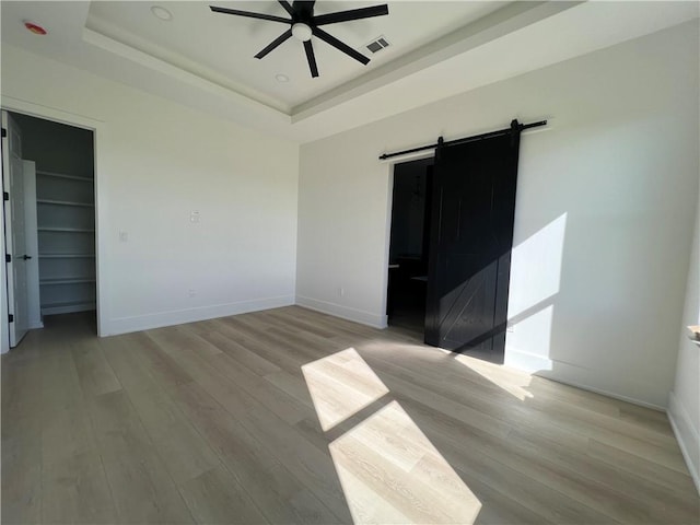 empty room with ceiling fan, a barn door, a raised ceiling, and light hardwood / wood-style flooring