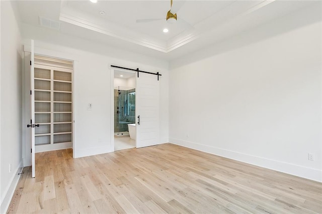 unfurnished bedroom featuring light hardwood / wood-style floors, ensuite bath, a barn door, ceiling fan, and a tray ceiling