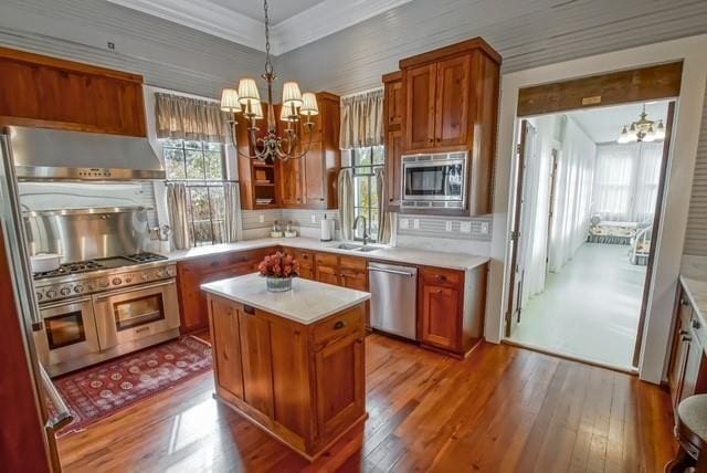 kitchen with pendant lighting, a chandelier, appliances with stainless steel finishes, wall chimney range hood, and hardwood / wood-style flooring