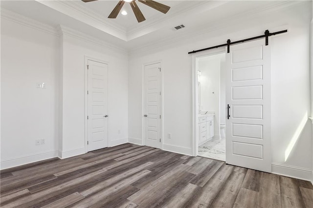 unfurnished bedroom with ceiling fan, ensuite bath, a barn door, dark hardwood / wood-style floors, and a tray ceiling