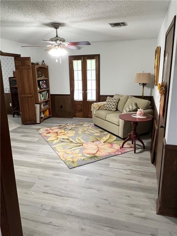 living room featuring french doors, wood walls, a textured ceiling, ceiling fan, and light hardwood / wood-style floors