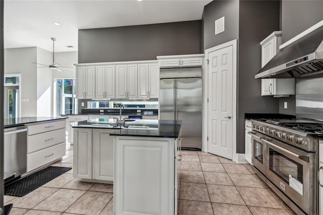 kitchen with white cabinetry, light tile floors, premium appliances, wall chimney exhaust hood, and ceiling fan