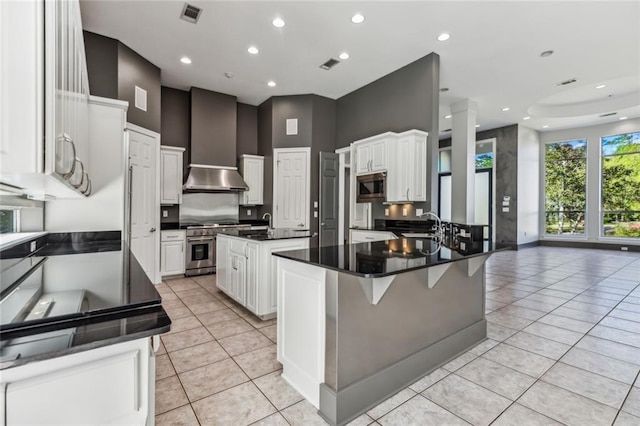 kitchen featuring appliances with stainless steel finishes, light tile flooring, a center island with sink, white cabinetry, and wall chimney exhaust hood