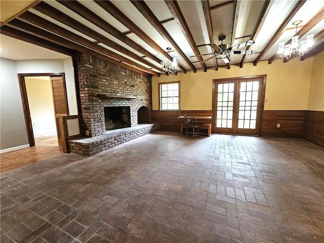 unfurnished living room featuring beamed ceiling, french doors, a brick fireplace, brick wall, and ceiling fan with notable chandelier