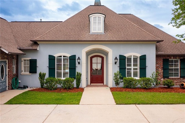 french country home featuring brick siding, stucco siding, a front yard, and roof with shingles