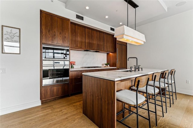 kitchen featuring sink, tasteful backsplash, decorative light fixtures, light wood-type flooring, and a kitchen island with sink