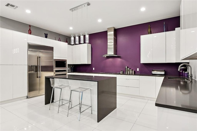 kitchen with wall chimney exhaust hood, built in appliances, hanging light fixtures, sink, and a kitchen island