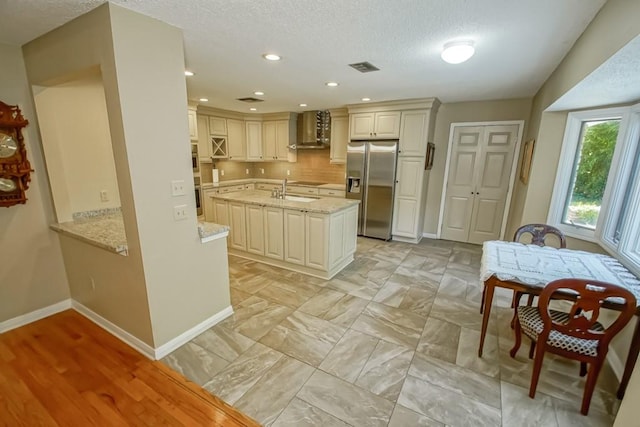 kitchen with light stone countertops, backsplash, stainless steel refrigerator with ice dispenser, wall chimney range hood, and light tile floors