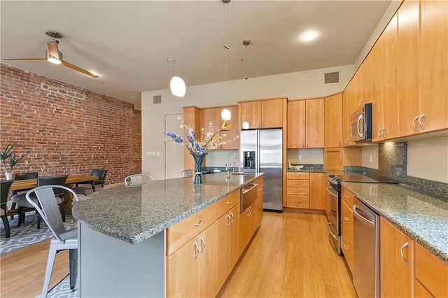 kitchen with brick wall, appliances with stainless steel finishes, an island with sink, and light hardwood / wood-style flooring