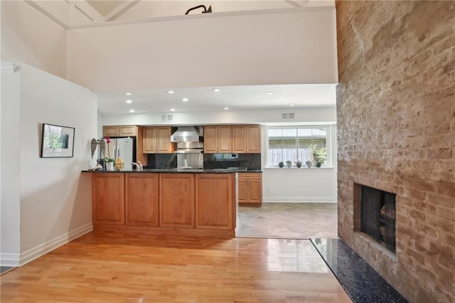 kitchen with stainless steel refrigerator, a large fireplace, wall chimney range hood, kitchen peninsula, and light wood-type flooring