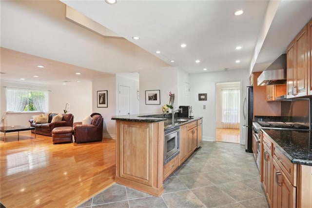 kitchen with wall chimney exhaust hood, stainless steel appliances, sink, dark stone countertops, and light hardwood / wood-style floors