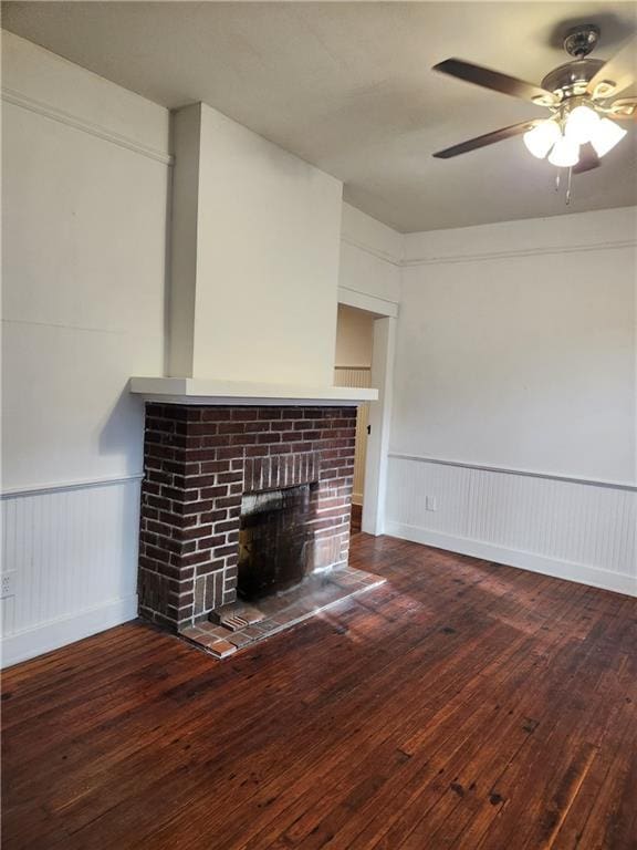unfurnished living room featuring a ceiling fan, a brick fireplace, hardwood / wood-style flooring, a wainscoted wall, and a decorative wall