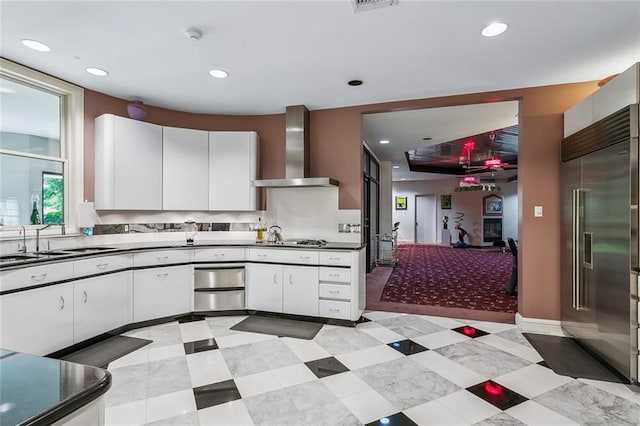 kitchen with dark countertops, wall chimney range hood, stainless steel built in refrigerator, white cabinetry, and a sink