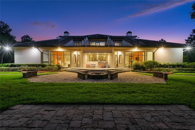 back of house at dusk with a lawn, a chimney, a patio, and stucco siding