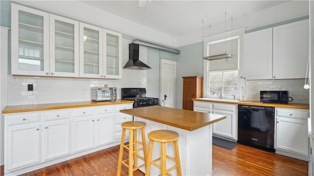 kitchen featuring white cabinetry, butcher block counters, a kitchen bar, black appliances, and wall chimney range hood