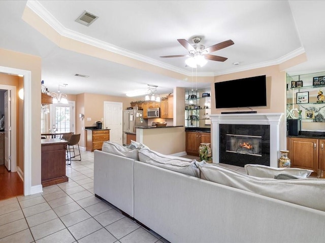 living room featuring ornamental molding, ceiling fan with notable chandelier, a raised ceiling, a tiled fireplace, and light tile patterned flooring