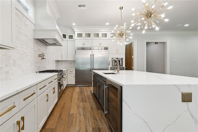 kitchen with an inviting chandelier, premium appliances, premium range hood, a center island with sink, and white cabinets