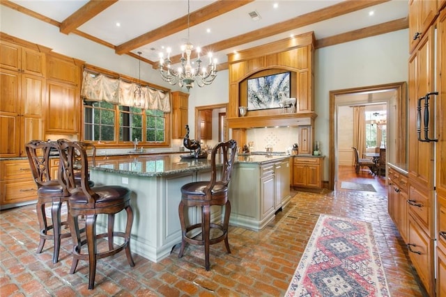 kitchen with backsplash, a center island with sink, dark stone counters, brick floor, and a notable chandelier