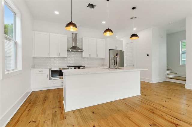 kitchen featuring light wood-type flooring, wall chimney range hood, backsplash, a kitchen island with sink, and pendant lighting