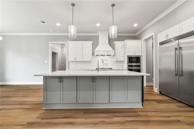 kitchen with built in appliances, light wood-type flooring, premium range hood, and an island with sink