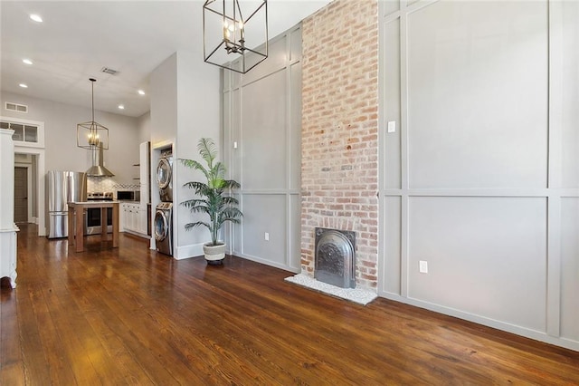 unfurnished living room with dark wood-type flooring, a chandelier, stacked washer and dryer, and a brick fireplace