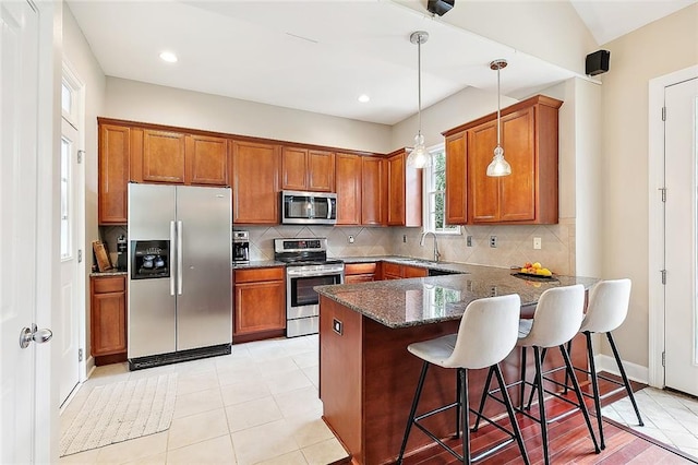 kitchen featuring kitchen peninsula, dark stone counters, appliances with stainless steel finishes, decorative light fixtures, and a kitchen bar