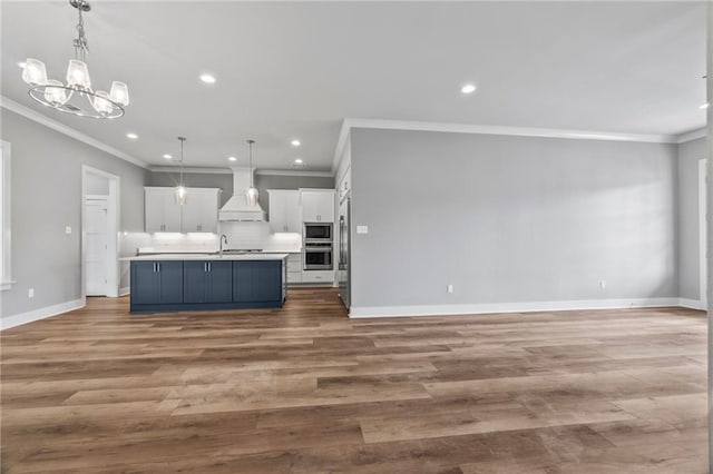 kitchen featuring appliances with stainless steel finishes, an island with sink, custom range hood, white cabinets, and decorative light fixtures