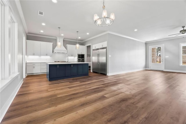 kitchen with ceiling fan with notable chandelier, a kitchen island with sink, stainless steel built in refrigerator, white cabinets, and decorative light fixtures