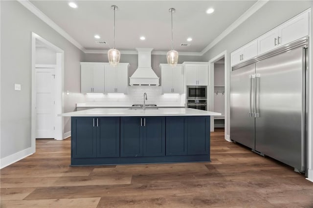 kitchen with white cabinetry, a kitchen island with sink, built in appliances, custom range hood, and decorative light fixtures