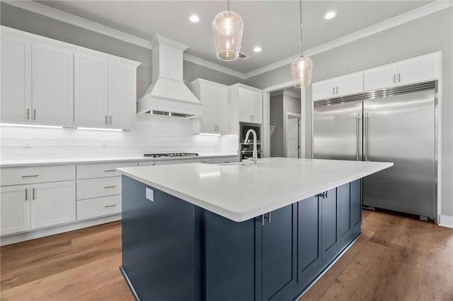 kitchen featuring built in fridge, decorative light fixtures, white cabinetry, custom range hood, and a center island with sink