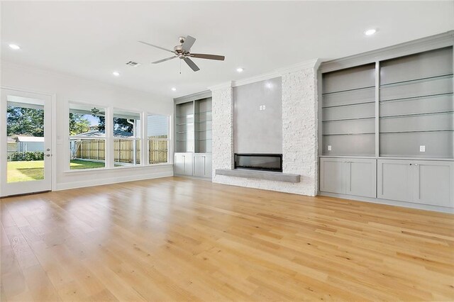 unfurnished living room with crown molding, built in shelves, a stone fireplace, ceiling fan, and light hardwood / wood-style flooring