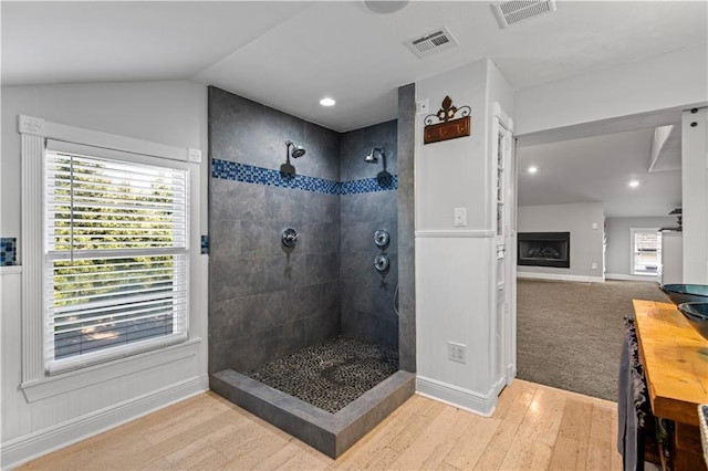 bathroom with wood-type flooring, a large fireplace, a tile shower, and vaulted ceiling