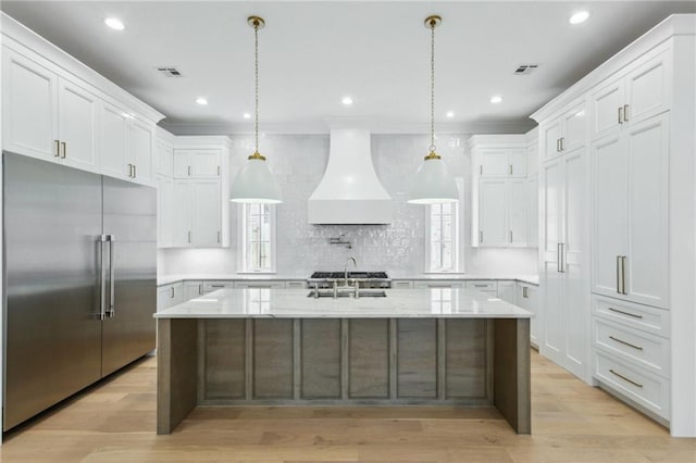 kitchen featuring a kitchen island with sink, built in fridge, custom range hood, and white cabinets