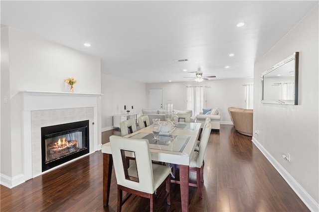 dining area with a tiled fireplace, dark wood-type flooring, and ceiling fan