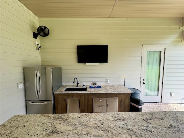 kitchen with stainless steel refrigerator, sink, light stone counters, wooden walls, and wooden ceiling