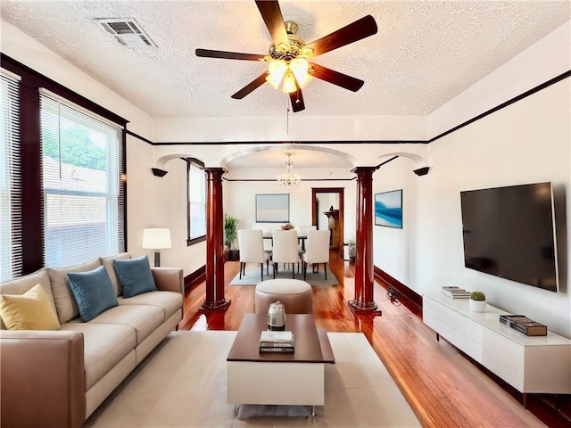 living room featuring ceiling fan with notable chandelier, light wood-type flooring, decorative columns, and a textured ceiling