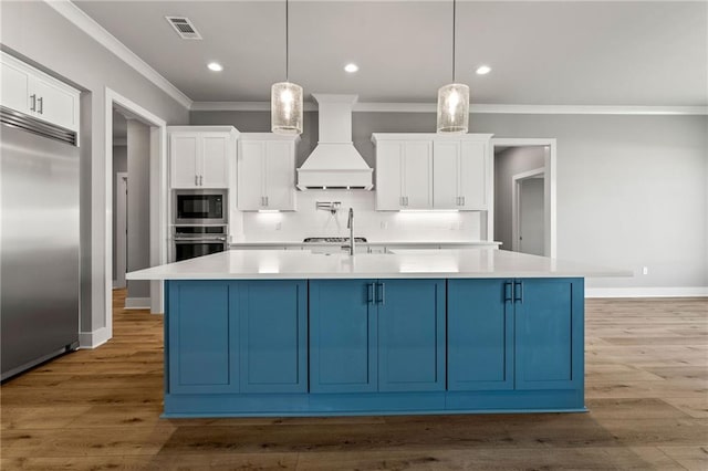 kitchen featuring custom range hood, built in appliances, pendant lighting, a center island with sink, and tasteful backsplash