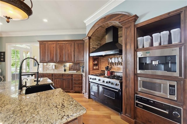 kitchen with sink, wall chimney exhaust hood, light stone counters, decorative backsplash, and appliances with stainless steel finishes