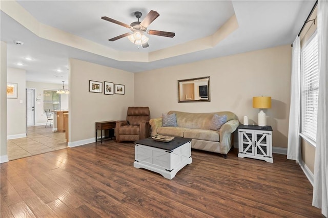 living area with a raised ceiling, ceiling fan with notable chandelier, dark wood-type flooring, and baseboards