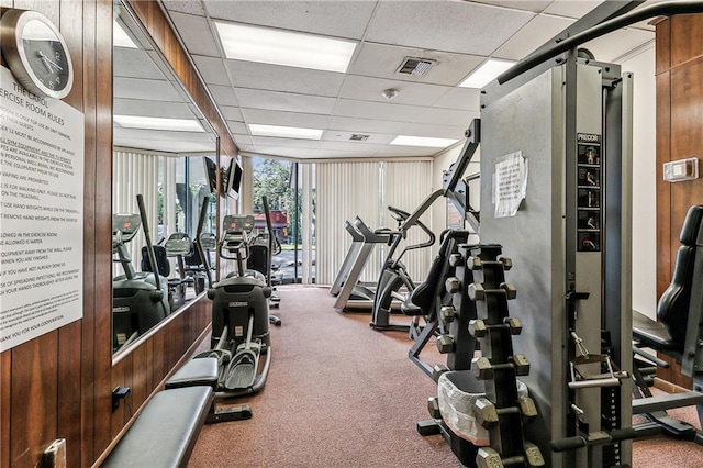 exercise room with carpet floors, a drop ceiling, and wooden walls