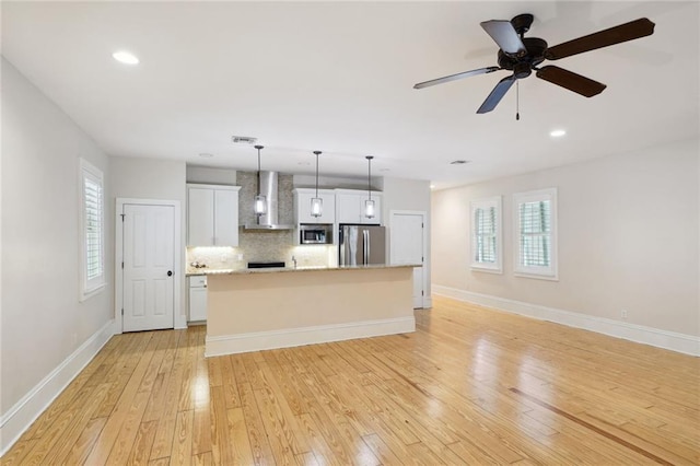 kitchen with hanging light fixtures, appliances with stainless steel finishes, white cabinetry, wall chimney range hood, and an island with sink