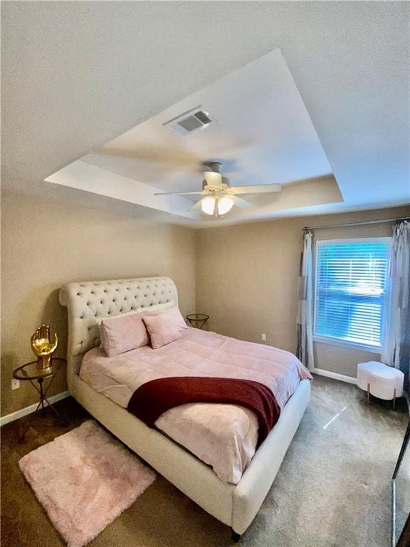 bedroom with a ceiling fan, visible vents, baseboards, dark colored carpet, and a tray ceiling