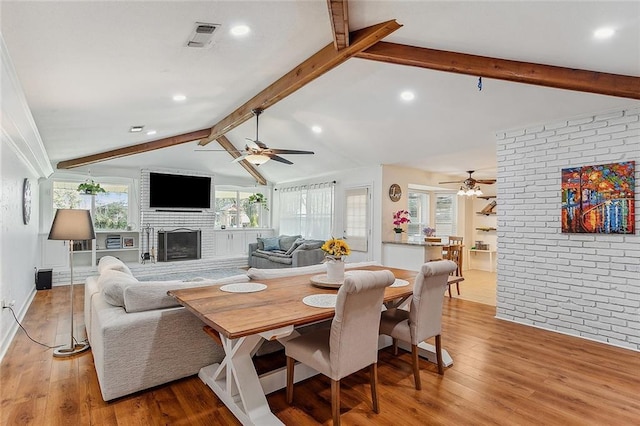 dining space featuring brick wall, light wood-type flooring, lofted ceiling with beams, and a fireplace