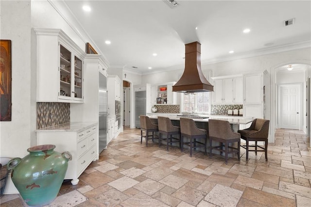 kitchen with arched walkways, glass insert cabinets, island exhaust hood, stone tile flooring, and white cabinetry