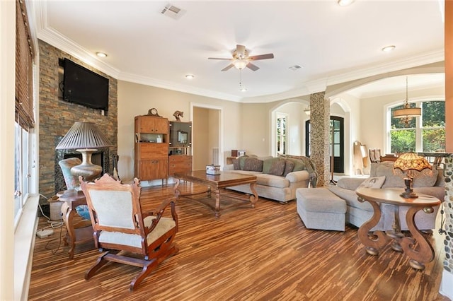 living room with a stone fireplace, decorative columns, wood-type flooring, ceiling fan, and crown molding