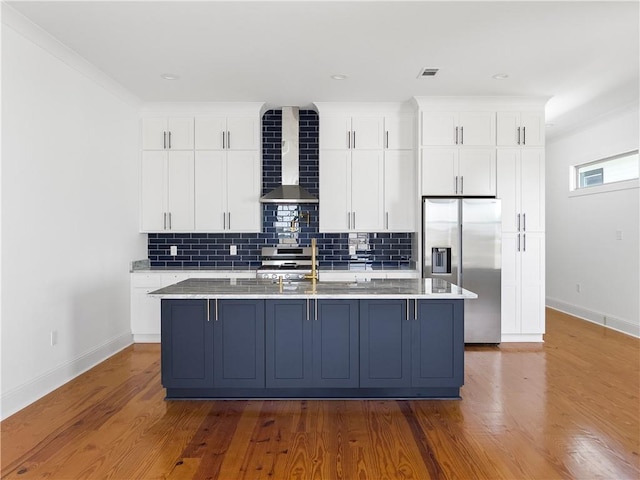 kitchen with baseboards, an island with sink, wall chimney exhaust hood, white cabinets, and appliances with stainless steel finishes