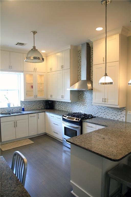 kitchen with white cabinetry, kitchen peninsula, wall chimney exhaust hood, stainless steel gas stove, and decorative light fixtures