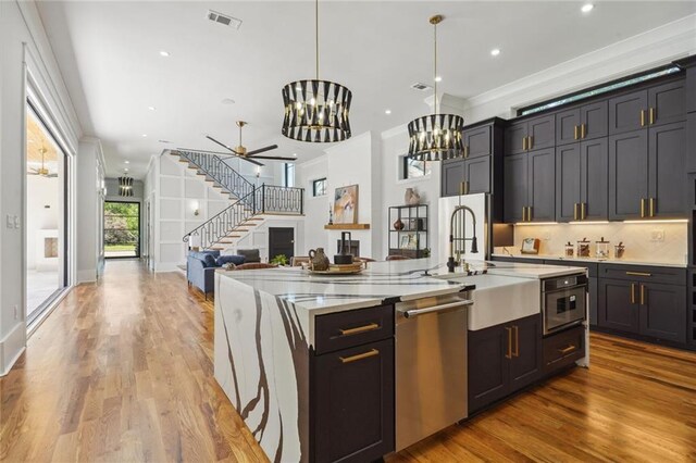 kitchen with crown molding, ceiling fan with notable chandelier, stainless steel appliances, wood-type flooring, and a kitchen island with sink