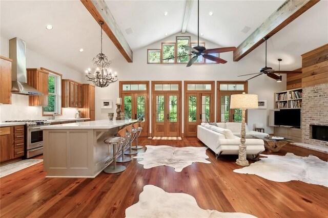 kitchen with stainless steel range with gas cooktop, ceiling fan with notable chandelier, a kitchen island, beam ceiling, and wall chimney range hood