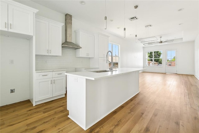 kitchen with hanging light fixtures, a kitchen island with sink, wall chimney range hood, and white cabinetry